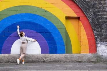 Young woman with hand raised leaning on rainbow flag painted on wall - AMWF00957