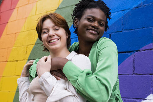 Happy woman embracing girlfriend in front of rainbow flag painted on wall - AMWF00960