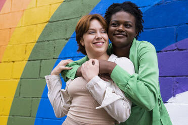 Smiling woman embracing girlfriend in front of rainbow flag painted on wall - AMWF00961