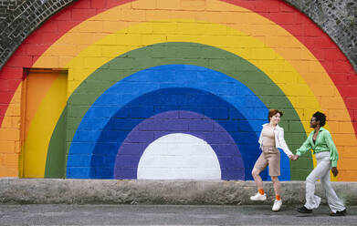 Happy lesbian couple holding hands walking on footpath in front of rainbow colored wall - AMWF00972