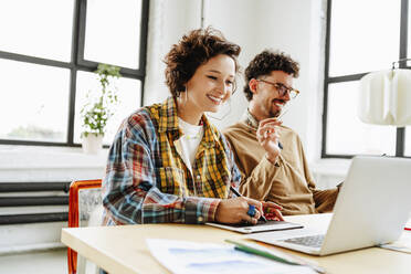 Happy graphic designer with colleague using laptop at desk in office - MDOF00054