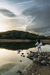 Astronaut holding space helmet standing on rock at beach - DAMF01104