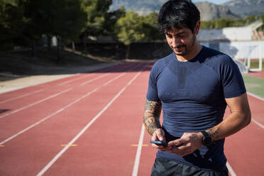 Muscular Hispanic male runner standing at stadium and browsing mobile phone during training in summer - ADSF39582