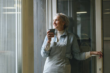 Smiling craftswoman holding coffee cup at doorway in workshop - YTF00282