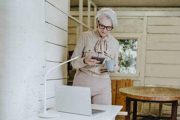 Reife Geschäftsfrau mit Kaffeetasse und Blick auf einen Tablet-PC vor einem Laptop auf dem Schreibtisch - YTF00312