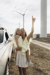 Smiling woman gesturing and standing with daughter by car at wind park - SIF00519