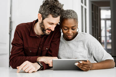 Zufriedene afroamerikanische Frau und bärtiger Mann lächelnd, während sie am Tisch sitzen und gemeinsam ein digitales Tablet zu Hause anschauen - ADSF39609