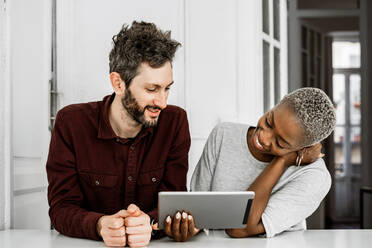 Zufriedene afroamerikanische Frau und bärtiger Mann lächelnd, während sie am Tisch sitzen und gemeinsam ein digitales Tablet zu Hause anschauen - ADSF39610