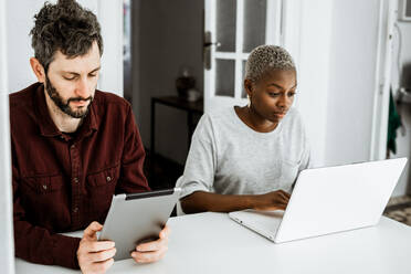 Seitenansicht einer zeitgenössischen afroamerikanischen Frau, die an einem Tisch sitzt und mit einem Laptop arbeitet, während ein junger Mann zu Hause auf einem digitalen Tisch surft - ADSF39611