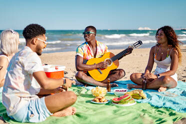 Multirassische Männer und Frauen in Freizeitkleidung trinken Bier und essen Früchte, während sie im Schneidersitz auf einer Decke sitzen, während eines Picknicks am Strand an einem Sommerwochenende - ADSF39622