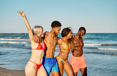 Happy multiracial men and women raising arms and embracing each other while strolling on beach near waving sea on summer weekend day - ADSF39625