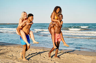 Seitenansicht von glücklichen multiethnischen Frauen, die auf den Rücken von schwarzen Freunden klettern, während sie an einem Sommertag in den schäumenden Meereswellen spielen - ADSF39626
