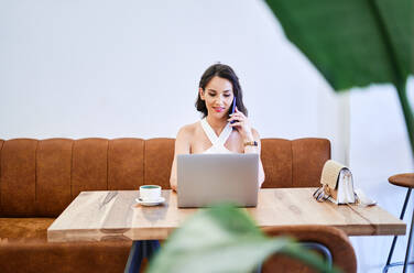 Eine Unternehmerin bespricht ein Geschäftsprojekt auf einem Mobiltelefon, während sie ein Netbook benutzt und aus der Ferne an einem Tisch in einem Café arbeitet - ADSF39630