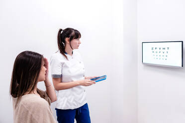 Optometristin, die die Sehkraft einer Patientin mit einer digitalen Testtafel während eines Besuchs in einer modernen Klinik untersucht - ADSF39641