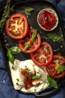 Von oben appetitlicher Burrata-Käse mit Sauce und frischen Tomatenscheiben mit Thymian und Basilikumblättern, serviert auf einem gusseisernen Tablett, das auf einer Tischdecke auf einem Holztisch steht - ADSF39674