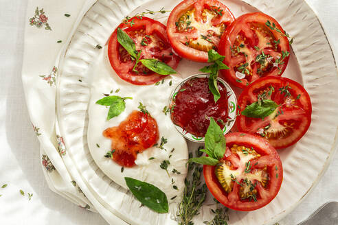 Draufsicht auf Scheiben frischer Tomaten und Mozzarella-Käse mit Soße und Kräutern, serviert auf einem Teller auf einem Tisch im Tageslicht - ADSF39675