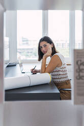 Smiling young businesswoman sitting with hand on chin in front of desktop - PNAF04597