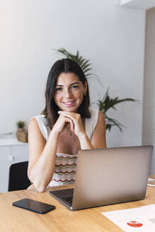 Glückliche junge Geschäftsfrau sitzt vor einem Laptop im Büro - PNAF04629