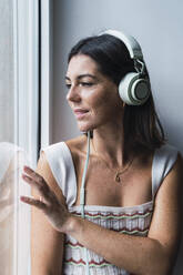 Nachdenkliche Geschäftsfrau mit Kopfhörern, die durch ein Fenster im Büro schaut - PNAF04641
