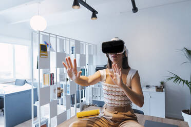 Businesswoman sitting on desk with VR goggles gesturing at work place - PNAF04642