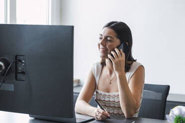 Lächelnde Geschäftsfrau, die über ihr Smartphone spricht und am Schreibtisch im Büro sitzt - PNAF04653
