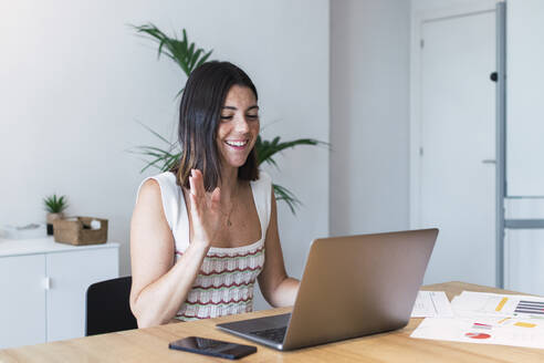 Lächelnde junge Geschäftsfrau bei einem Videoanruf, die mit ihrem Laptop winkt - PNAF04660