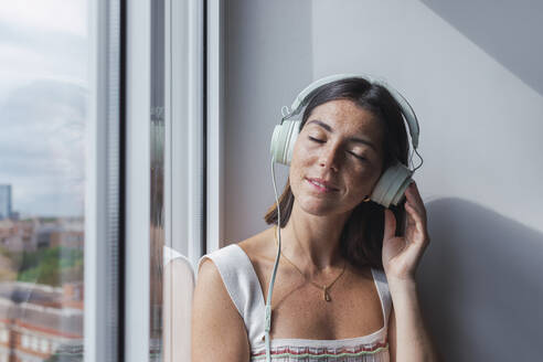 Junge Geschäftsfrau, die über Kopfhörer am Fenster Musik hört - PNAF04665