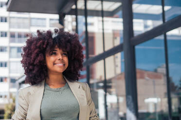 Positive black businesswoman in suit with curly hair standing looking away on modern city street before work - ADSF39676