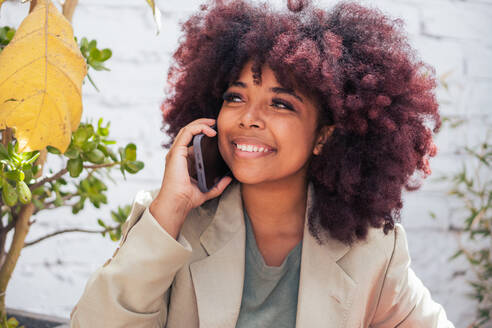 Fröhliche afroamerikanische Managerin, die lächelt, wegschaut und mit der Hand winkt, während sie einen Videoanruf über ein Handy auf der Straße tätigt - ADSF39677
