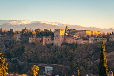 Atemberaubender Blick auf die sonnenbeschienene Festungsanlage Alhambra auf einem üppigen Hügel in der Nähe einer Bergkette bei Sonnenaufgang in Granada, Spanien - ADSF39681