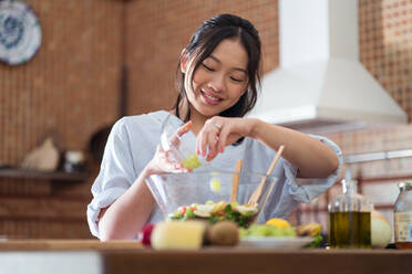 Asiatische Frau, die reife Trauben in eine Schüssel mit leckerem, gesundem Salat legt, während sie tagsüber zu Hause ihr Mittagessen zubereitet - ADSF39692