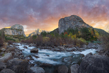 Schneller Strom sauberen Wassers, der auf Felsen in der Nähe eines mit Waldbäumen bewachsenen Bergrückens fließt, während eines Herbsttages mit Sonnenuntergang im Nationalpark Ordesa y Monte Perdido in Huesca, Spanien - ADSF39696