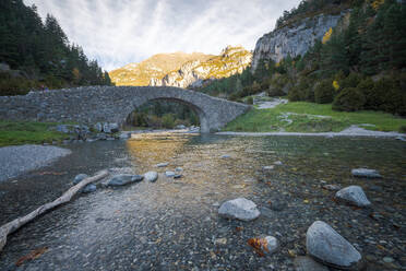 Schneller Fluss mit transparentem Wasser, der unter einer Steinbrücke gegen eine Bergspitze und einen bewölkten Himmel an einem sonnigen Tag im Nationalpark Ordesa y Monte Perdido in Huesca, Spanien, fließt - ADSF39700