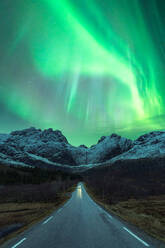 Scenic view of automobile driving on empty mountainous road in winter under night sky glowing with bright green aurora borealis in Norway - ADSF39732