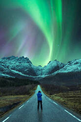 Distant view of explorer standing on asphalt road in snowy mountains under dark night sky with bright green polar lights in Norway - ADSF39733