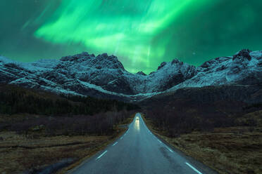 Landschaftlicher Blick auf ein Auto, das auf einer leeren Bergstraße im Winter fährt, während der Nachthimmel mit hellen grünen Polarlichtern in Norwegen leuchtet - ADSF39734