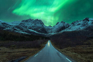 Scenic view of automobile driving on empty mountainous road in winter under night sky glowing with bright green aurora borealis in Norway - ADSF39734