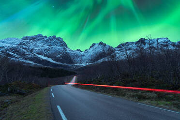 Atemberaubende Landschaft von felsigen Bergkette mit Schnee und leeren Asphaltstraße unter Nachthimmel mit lila und grünen Polarlichtern in Norwegen bedeckt - ADSF39735