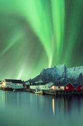 Spektakulärer Blick auf lebendige grüne Polarlichter, die am Himmel über Wohnhäusern im Hochland in der Nähe eines Sees in Norwegen bei Nacht leuchten - ADSF39737
