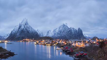 Spektakuläre Kulisse typischer Wohnhäuser am Ufer des ruhigen Meeres, umgeben von massiven schneebedeckten Felsenbergen unter nebligem Himmel auf den Lofoten - ADSF39739