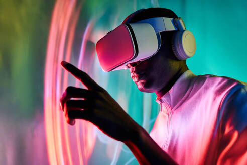 Seitenansicht eines konzentrierten jungen Afroamerikaners in weißem T-Shirt und moderner VR-Brille, der auf den Bildschirm zeigt und ihn berührt, während er mit einem virtuellen Objekt in einem dunklen Studio mit neonfarbener Beleuchtung interagiert - ADSF39751