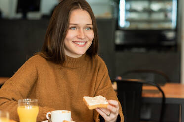 Content young female with long hair in warm sweater smiling and looking away while eating delicious toast sitting at table with glass of juice and cup of coffee in cafe - ADSF39755