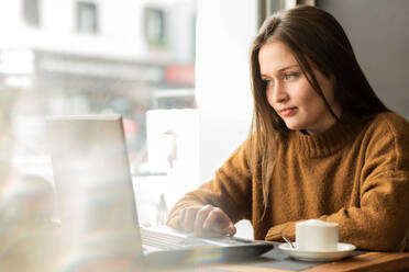 Nachdenkliche junge Freiberuflerin im warmen Pullover mit langen dunklen Haaren, die nachdenklich wegschaut, während sie mit Laptop und einer Tasse Cappuccino in einem Café sitzt - ADSF39757