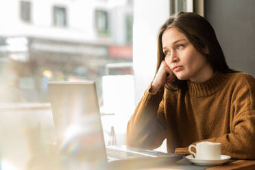 Thoughtful young female freelancer in warm sweater with long dark hair leaning on hand and looking away pensively while sitting in cafe with laptop and cup of cappuccino - ADSF39758