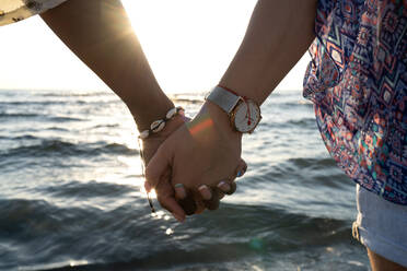 Anonyme verschiedene Freundinnen mit Armband und Armbanduhr halten Hände gegen wogende Meer und Sonnenuntergang Himmel während romantisches Date am Strand - ADSF39759