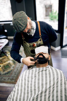 Konzentrierter unrasierter männlicher Friseur mittleren Alters in Schürze und Baskenmütze, der den dunklen Bart eines ethnischen Kunden mit einer Schere in einem traditionellen Friseursalon schneidet - ADSF39762