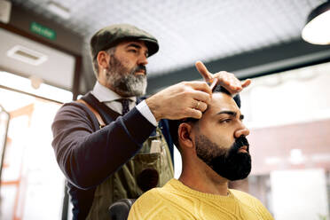 Männlicher Friseur mit Kamm bürstet das Haar eines ethnischen hispanischen Kunden, während er eine Frisur in einem modernen Friseursalon macht - ADSF39764