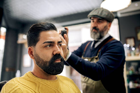 Konzentrierter bärtiger ethnischer Barbier mittleren Alters in Schürze und Baskenmütze, der Stylingprodukte auf das dunkle Haar eines ernsthaften Kunden in einem Schönheitssalon sprüht - ADSF39765