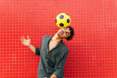 Young Hispanic man in black shirt balancing yellow football ball on head against tiled red wall on street - ADSF39809