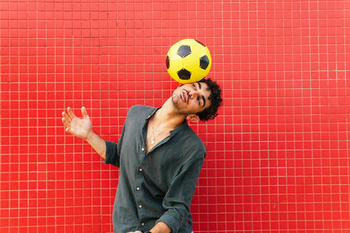 Junger hispanischer Mann in schwarzem Hemd balanciert gelben Fußball auf dem Kopf gegen eine rote Kachelwand auf der Straße - ADSF39809